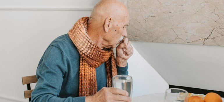 Deda koji kašlje pa je stavio ruku na usta dok u jednoj ruci drži čašu vode.