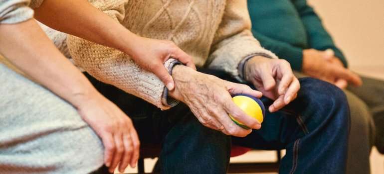 Starija osoba koju pomaže negovatelj, dok drži terapijsku lopticu u ruci.