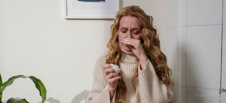 Женщина с длинными светлыми волосами вытирает нос, явно страдая от неприятных симптомов аллергического ринита.